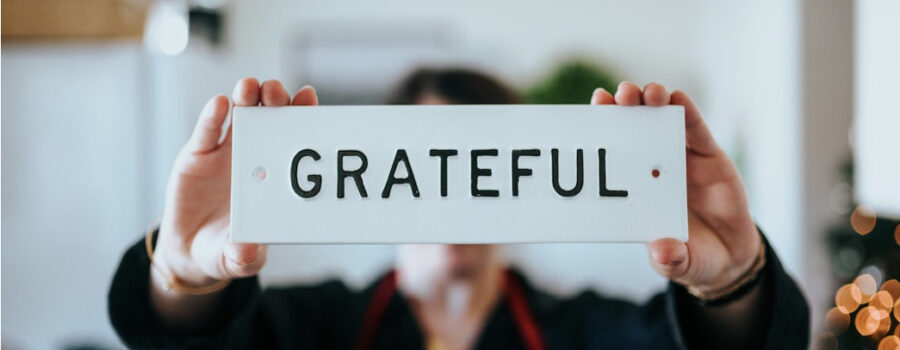 Woman holding a small sign that says "GRATEFUL". This photo is on www.becomingstoic.net in a post called "Always Be Grateful".