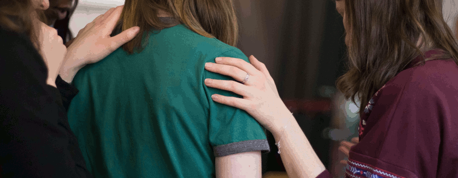 Three women surround a woman wearing a green shirt. They are laying their hands on her shoulder to show support. This photo is on www.becomingstoic.net in a post called "Devotion and Duty".