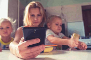 A mother is holding an infant in her left arms as it is turning over a glass. The mother holds a cellphone in her right hand and is watching it intently. Another small child is to her right. This photo is used in www.becomingstoic.net in an article called "Dealing with Distractions".